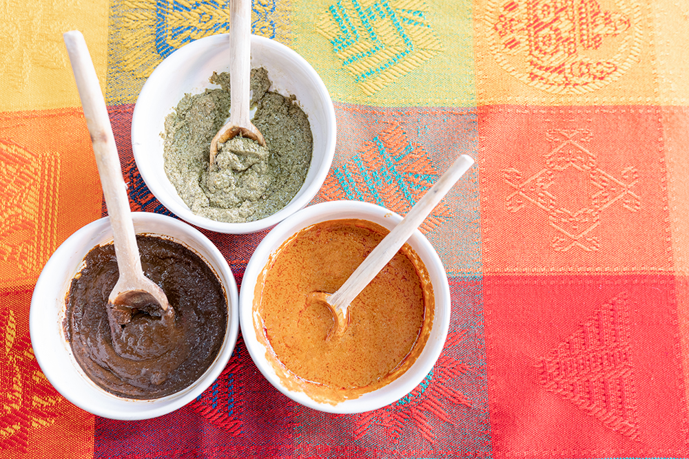 Three bowls with red, green and yellow mole sauce with a wooden spoon on top of a colorful traditional tablecloth. Concept of traditional Mexican food from Puebla and Oaxaca